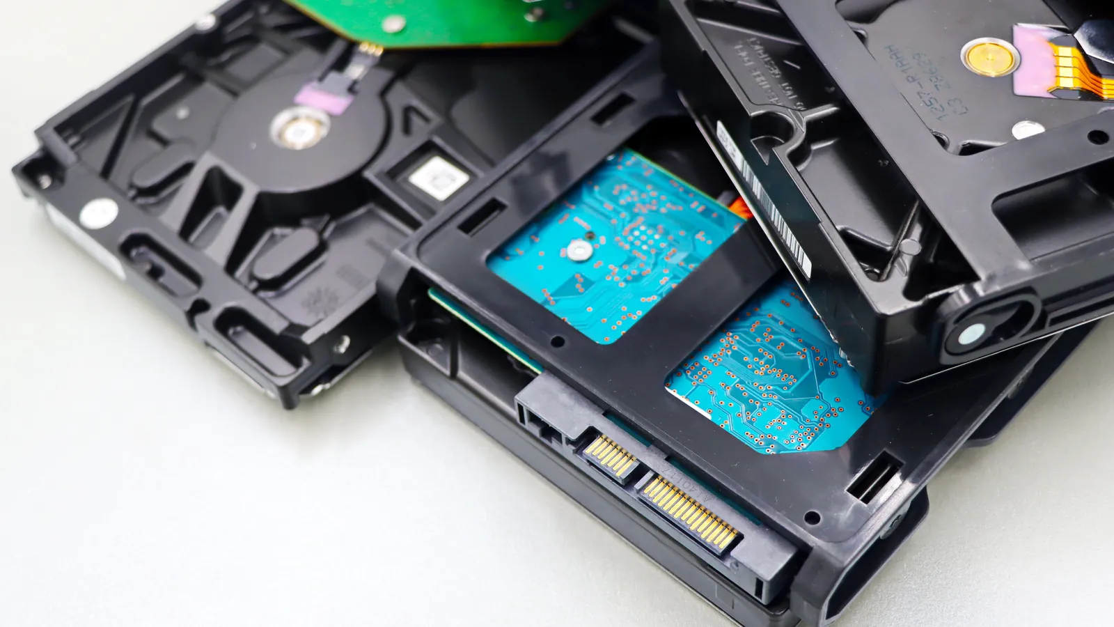 A stack of black internal hard disk drives with exposed teal circuit boards and gold connectors lying on a light grey surface.
