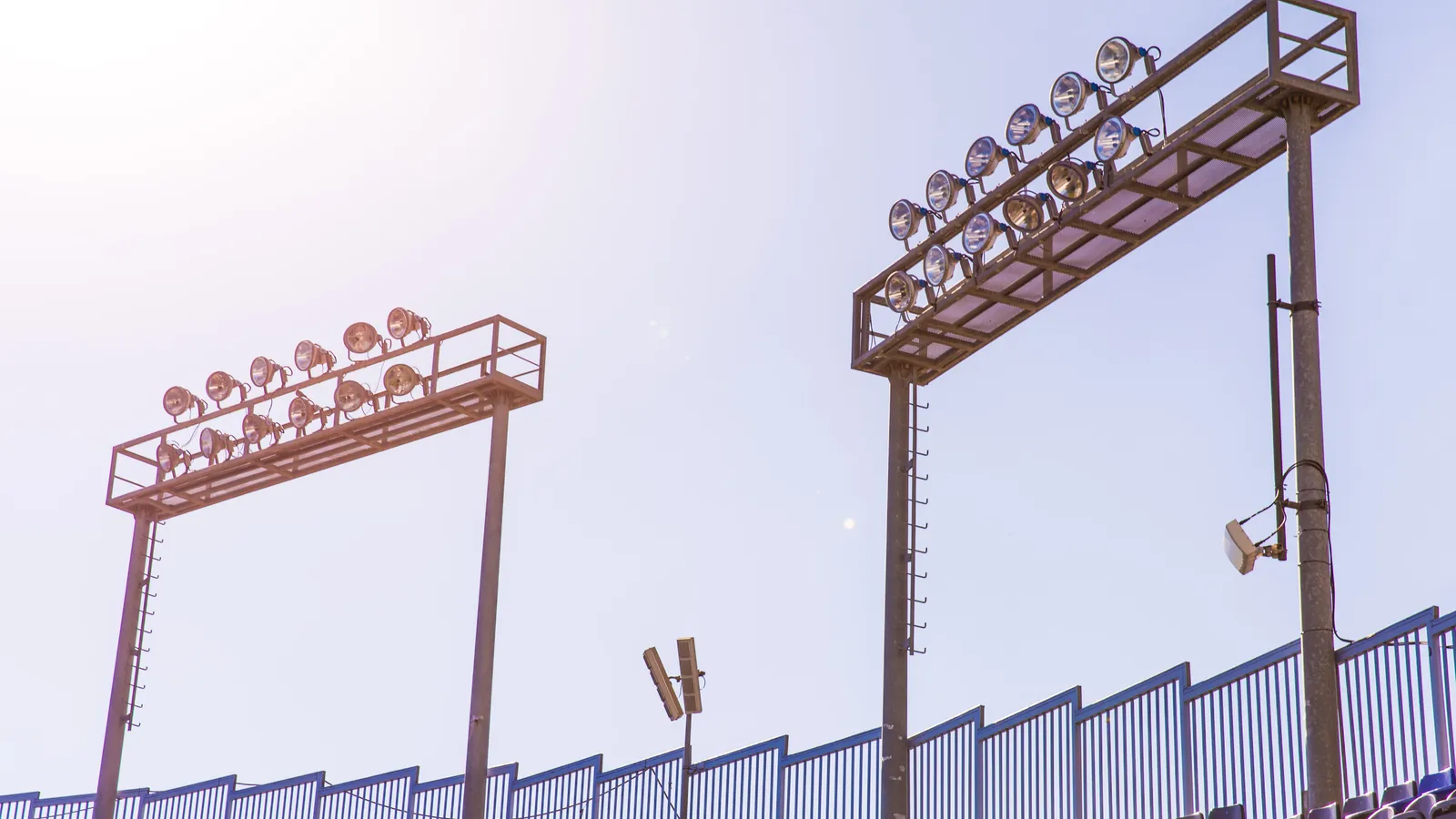 Two banks of stadium floodlights on metal frames against a pale blue sky, above rows of empty blue seats.