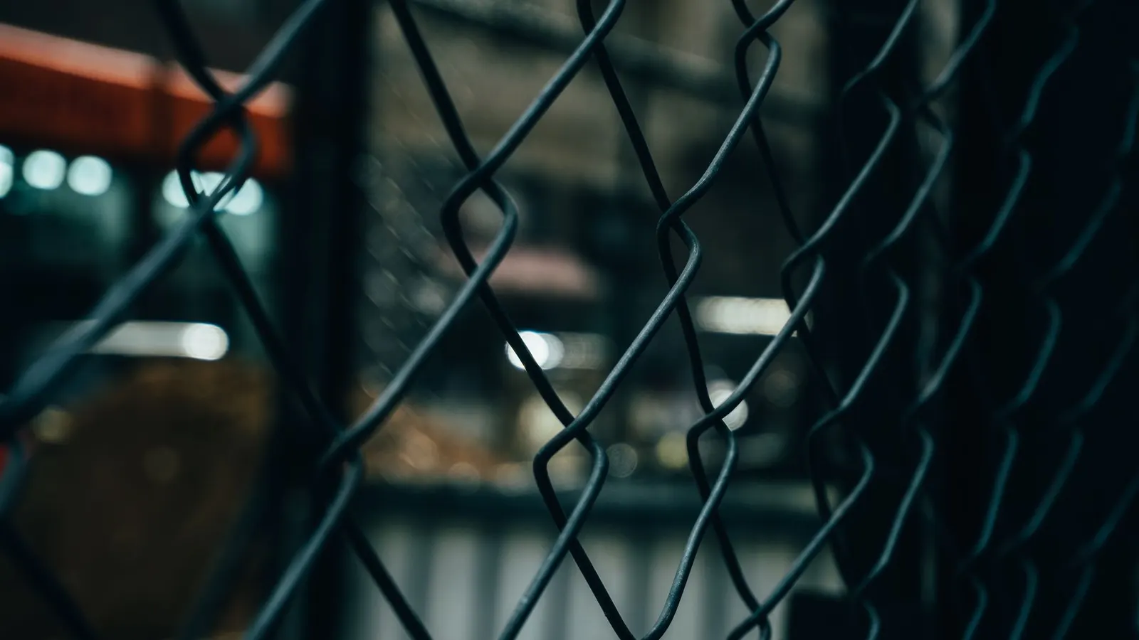 Close-up of a dark chain-link fence at night, with blurred orange and white lights behind it.