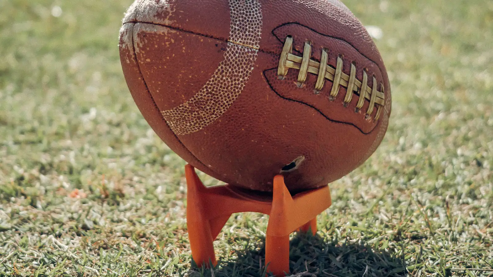 A worn American football standing on an orange kicking tee in sunlit grass.