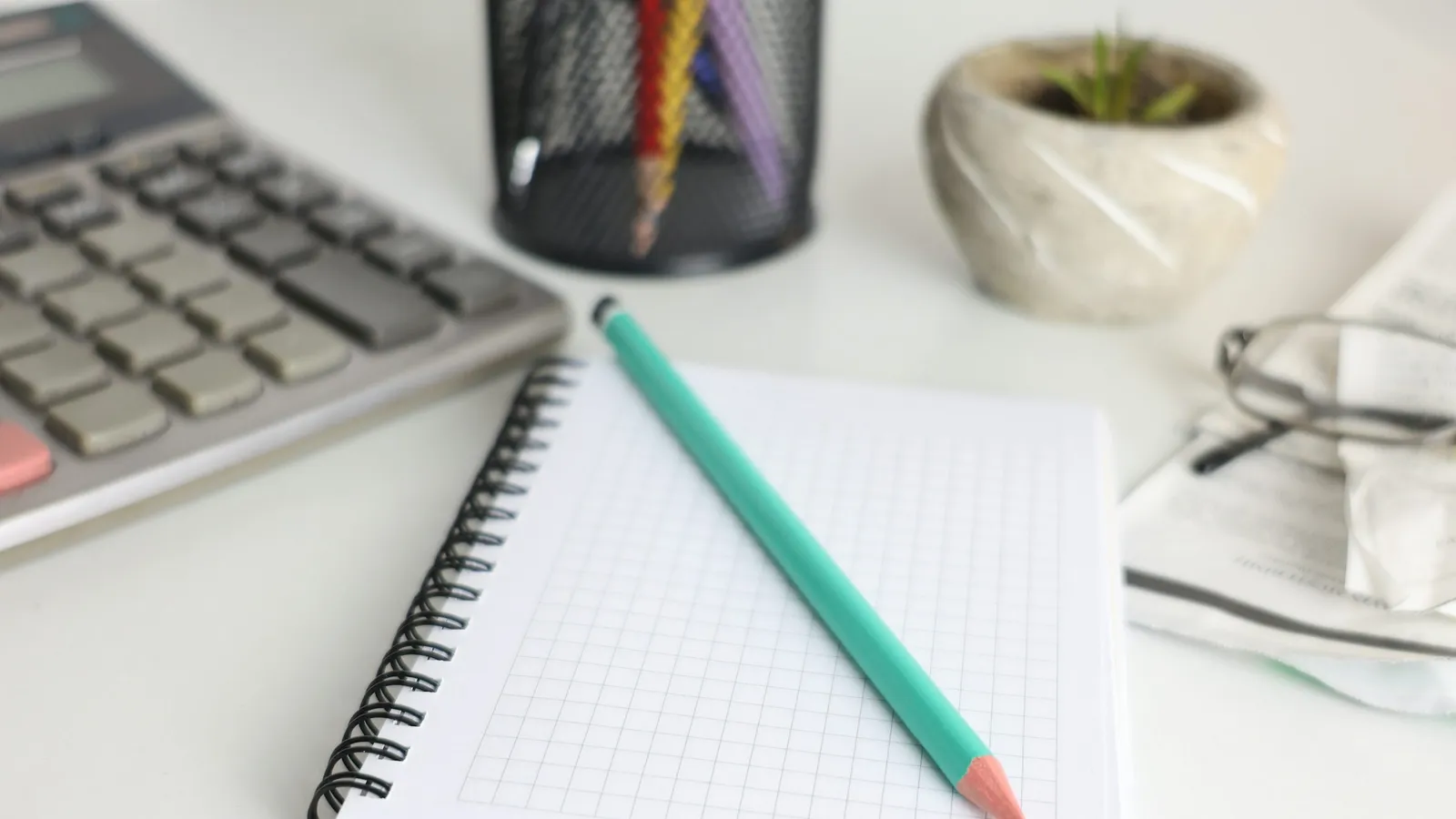 A calculator, grid notebook and green pencil beside reading glasses and a plant on a desk