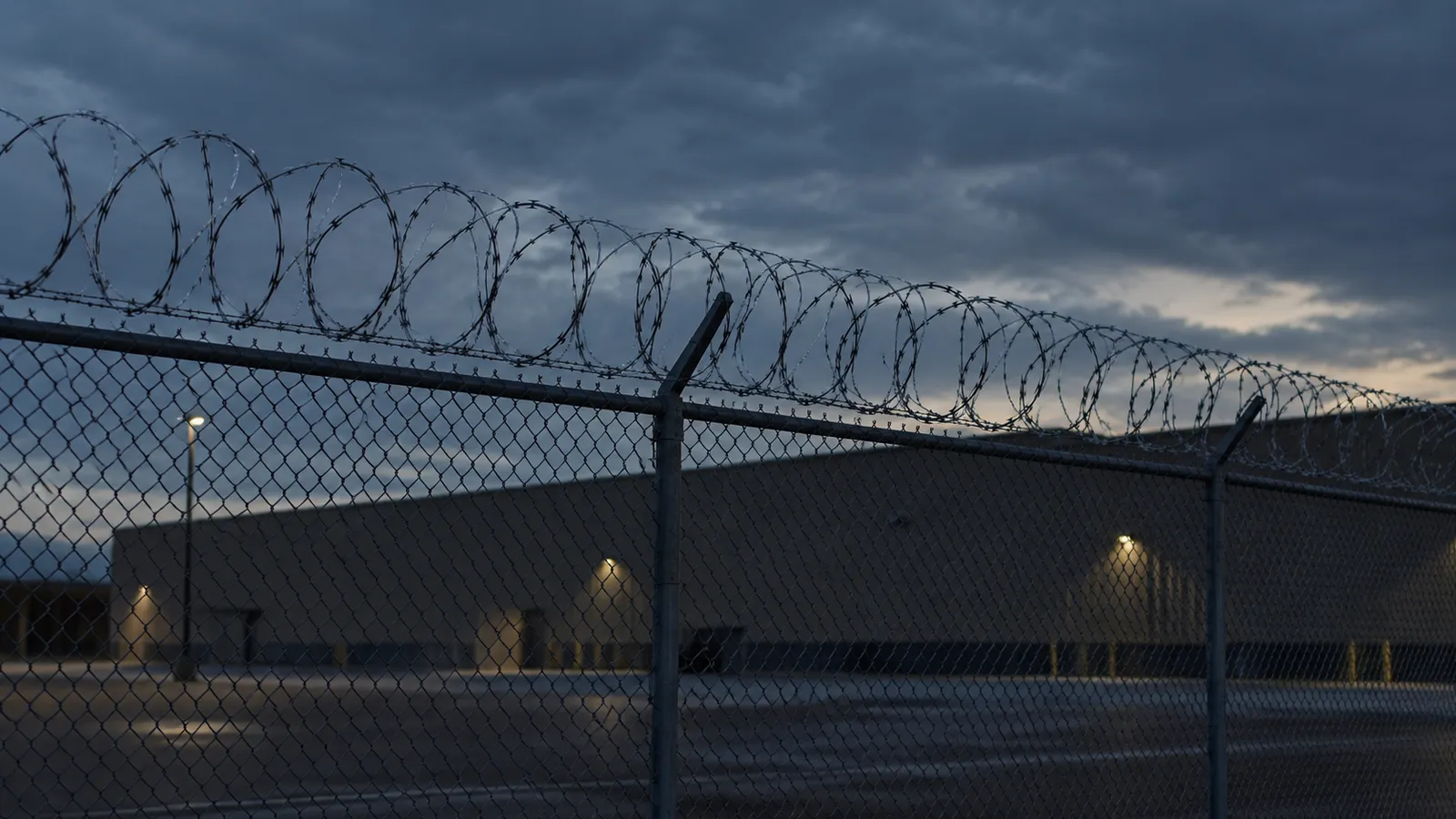 Coils of razor wire along the top of a tall security fence at dusk, with a low windowless building behind it