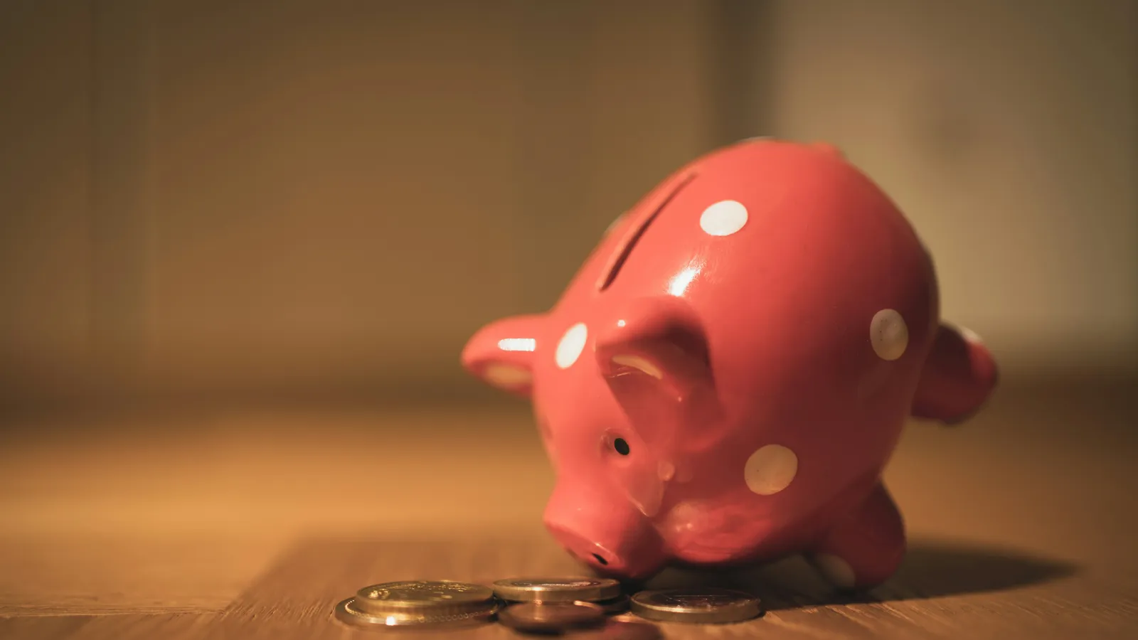 A red piggy bank with white polka dots nosing a small pile of coins on a wooden table